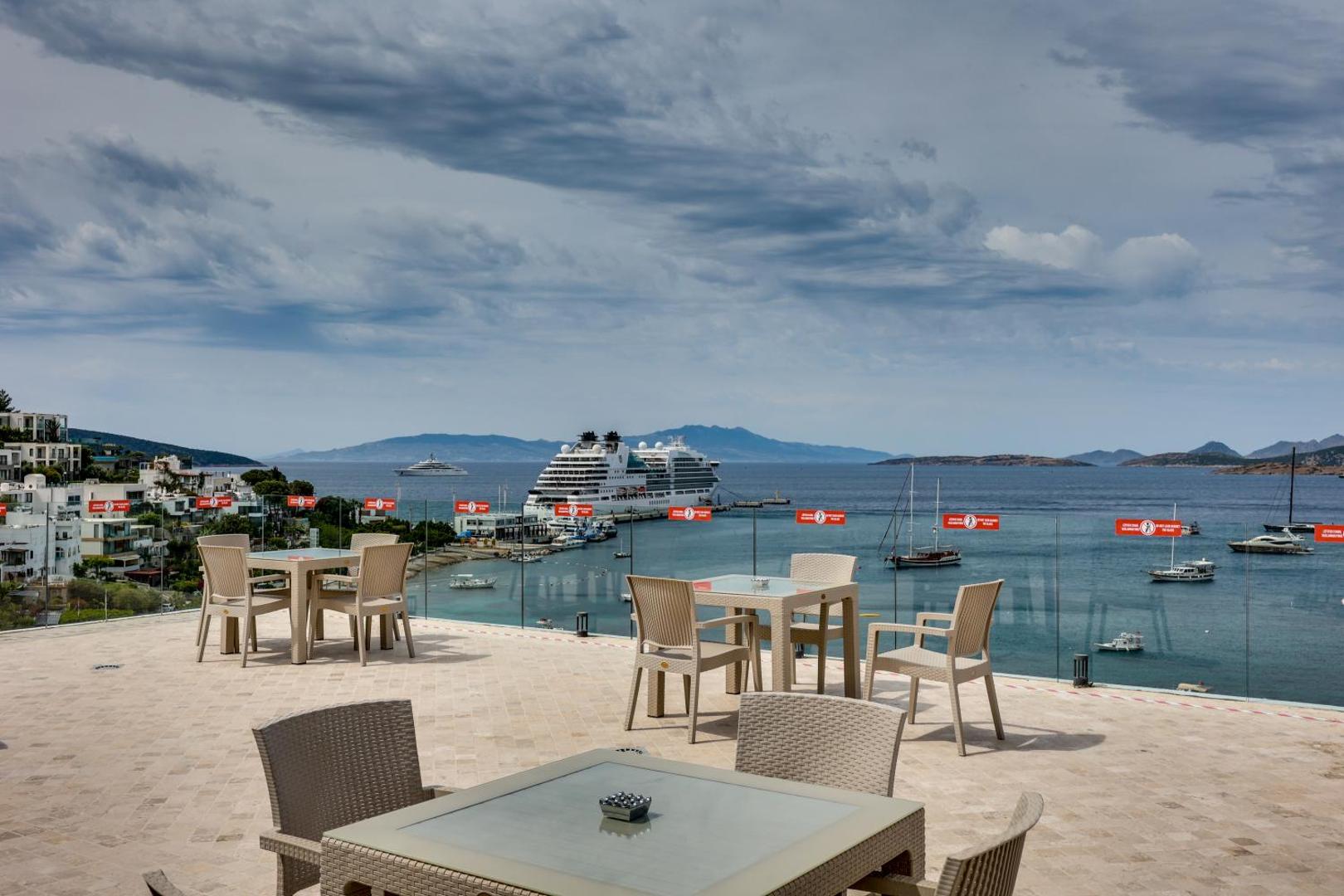 Diamond of Bodrum Hotel āra terase ar galdiem un krēsliem, no kuras paveras skats uz līci un lielu kruīza kuģi