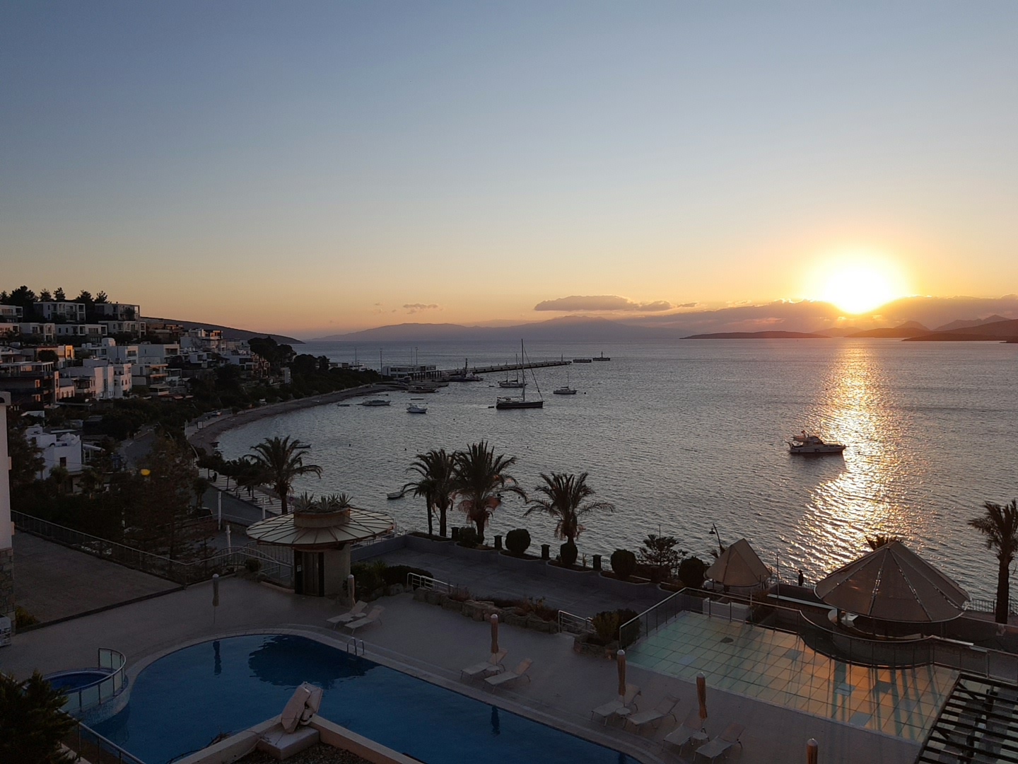 Saulriets virs Egejas jūras no Diamond of Bodrum Hotel terases ar baseinu, palmām un piekrastes apbūvi vakara saules gaismā