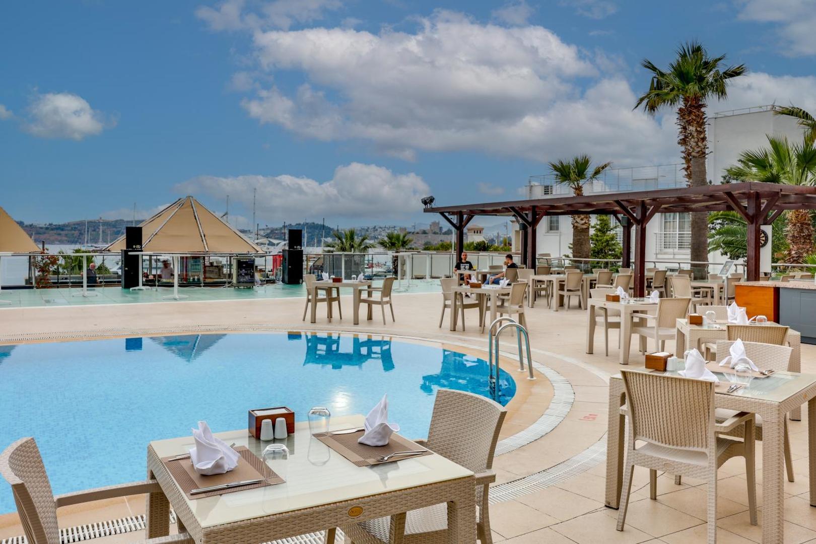 Diamond of Bodrum Hotel āra restorāna zona pie baseina ar klātiem galdiem, palmām un skatu uz jūru dienas laikā.