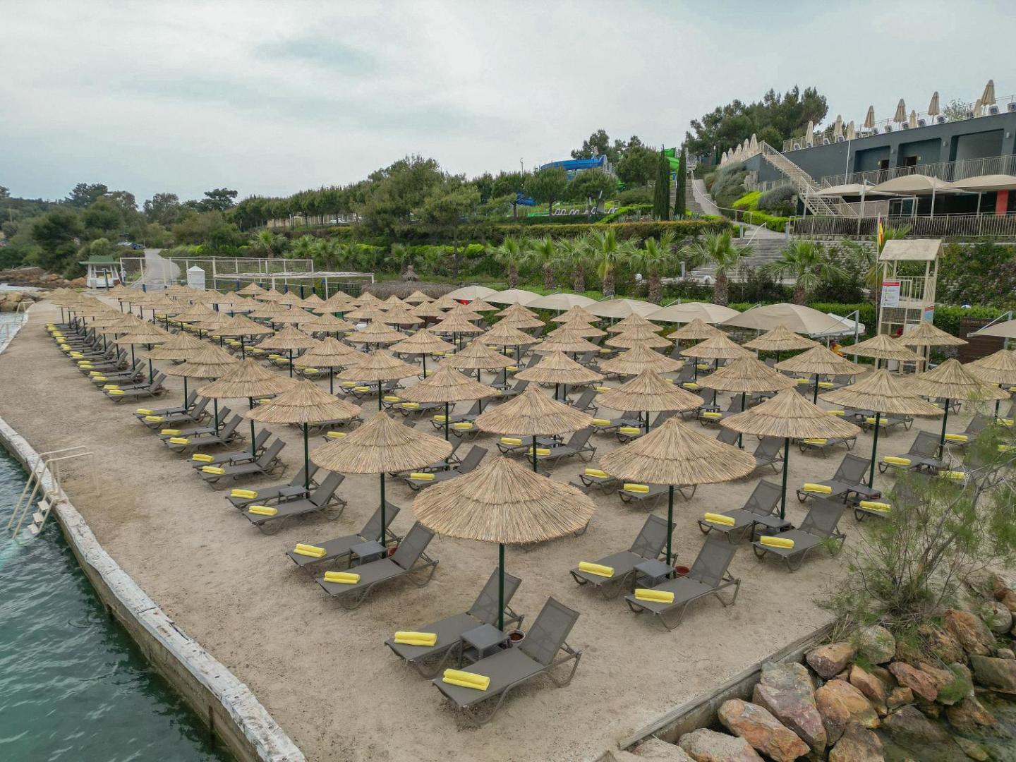 La Blanche Island Bodrum plašā pludmales zona ar salmu saulessargiem un ērtiem zviļņiem, fonā redzama viesnīcas teritorija un apstādījumi