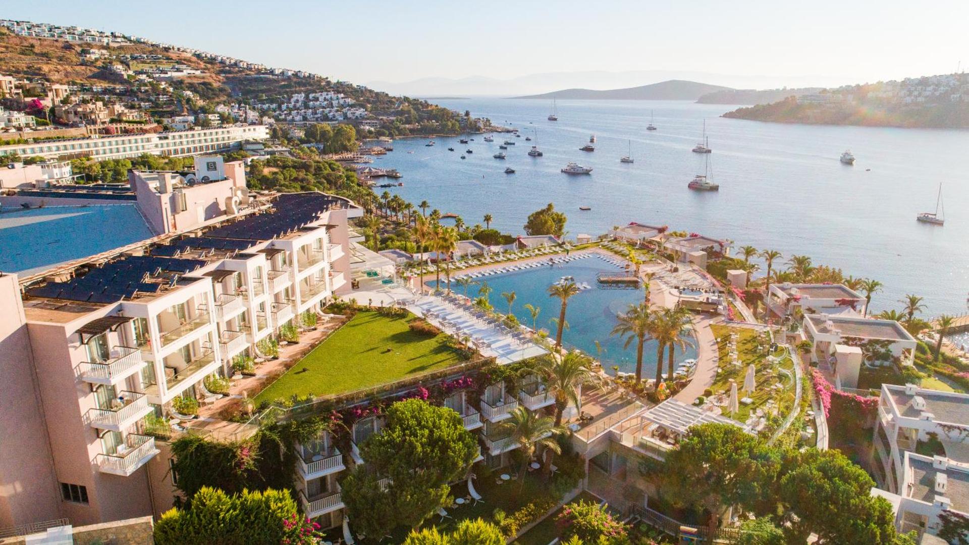 Skats no Hotel Baia Bodrum terases uz baseinu, piekrasti un jūrā noenkurotām jahtām saulrieta laikā Bodrumā.