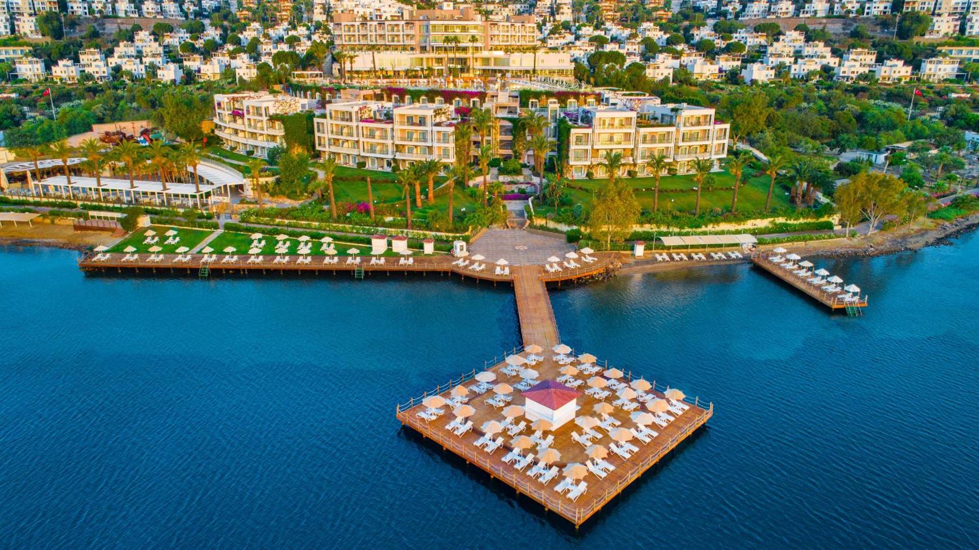 Hotel Baia Bodrum skats no jūras puses uz viesnīcas ēkām, zaļo teritoriju un koka laipu ar sauļošanās platformu Bodrumas piekrastē.