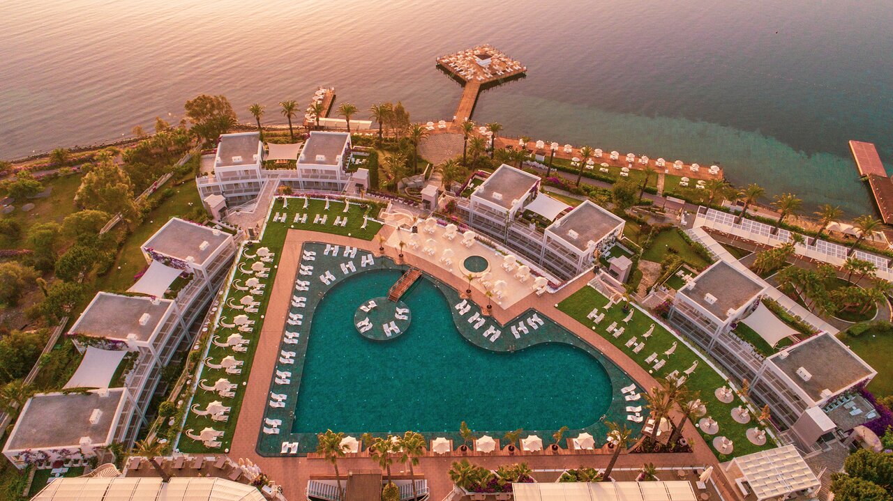 Aerofoto ar Hotel Baia Bodrum sirds formas baseinu, privāto piestātni un viesnīcas korpusiem saulrieta gaismā pie jūras