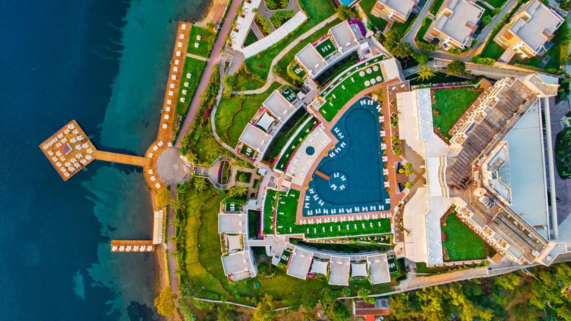 Hotel Baia Bodrum teritorijas skats no putna lidojuma, parādot baseina formu, dārzus, ēku izvietojumu un pieeju jūrai.