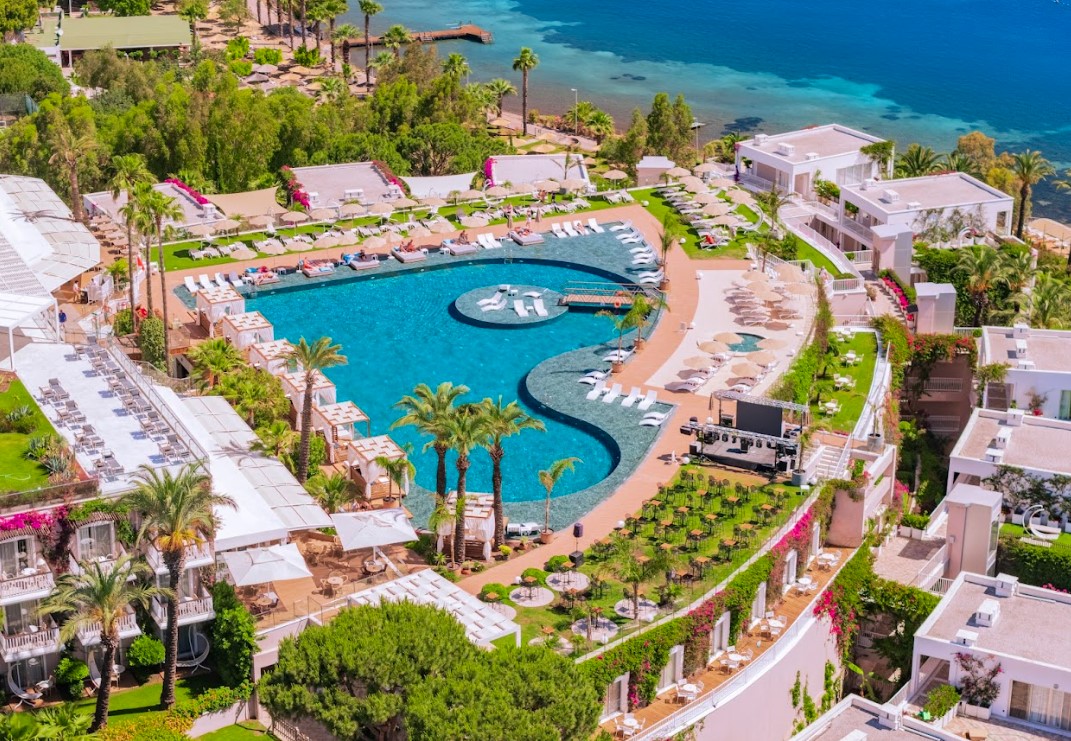 Hotel Baia Bodrum teritorijas skats no augšas ar sirds formas baseinu, dārzu, bungalo tipa ēkām un jūras krastu
