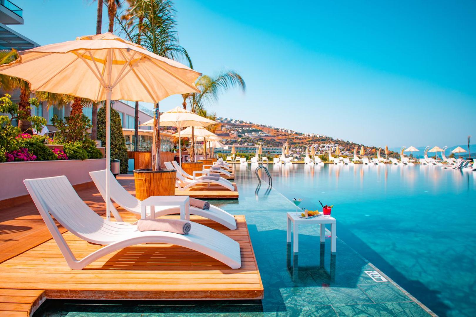 Hotel Baia Bodrum sauļošanās zona ar baltiem zviļņiem uz koka terases pie dzidra baseina saulainā dienā
