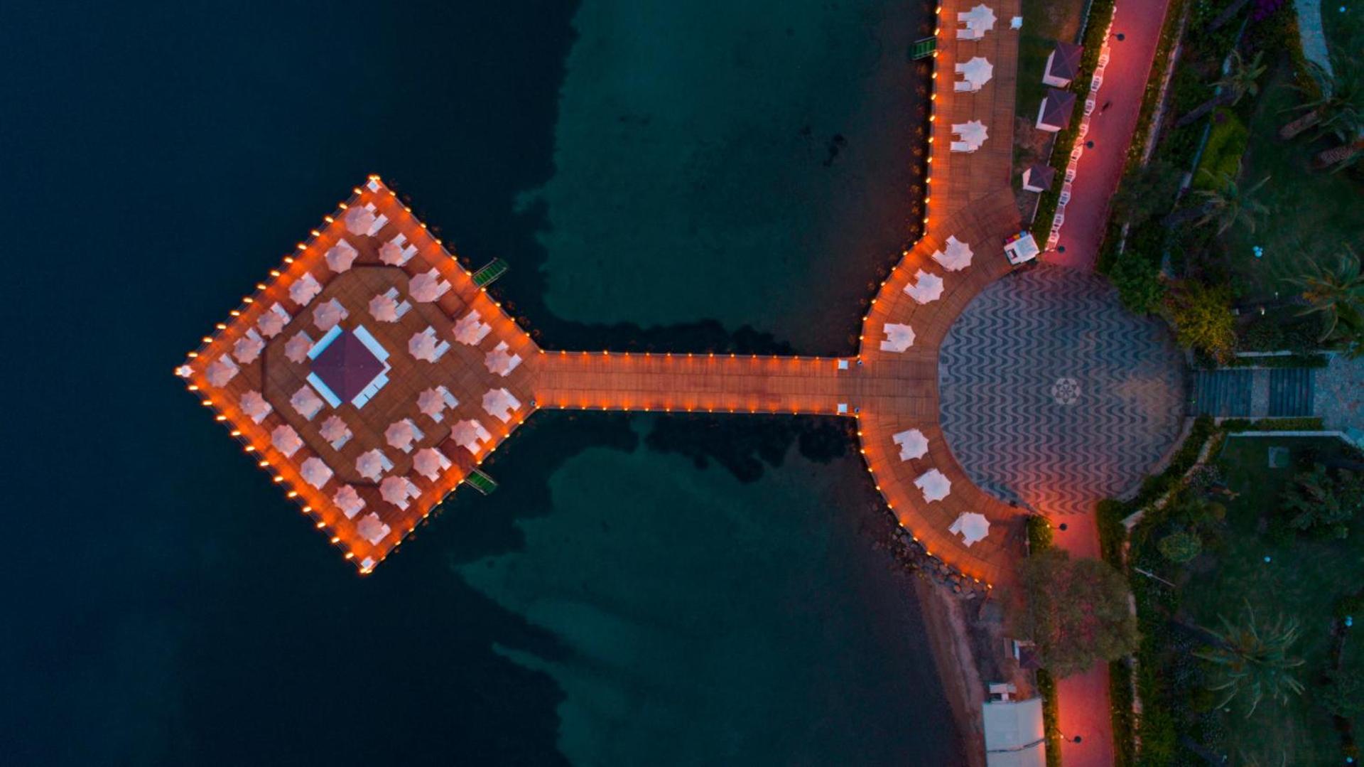 Vertikāls aerofoto skats uz Hotel Baia Bodrum izgaismoto piestātni naktī ar simetriski izvietotiem sauļošanās krēsliem