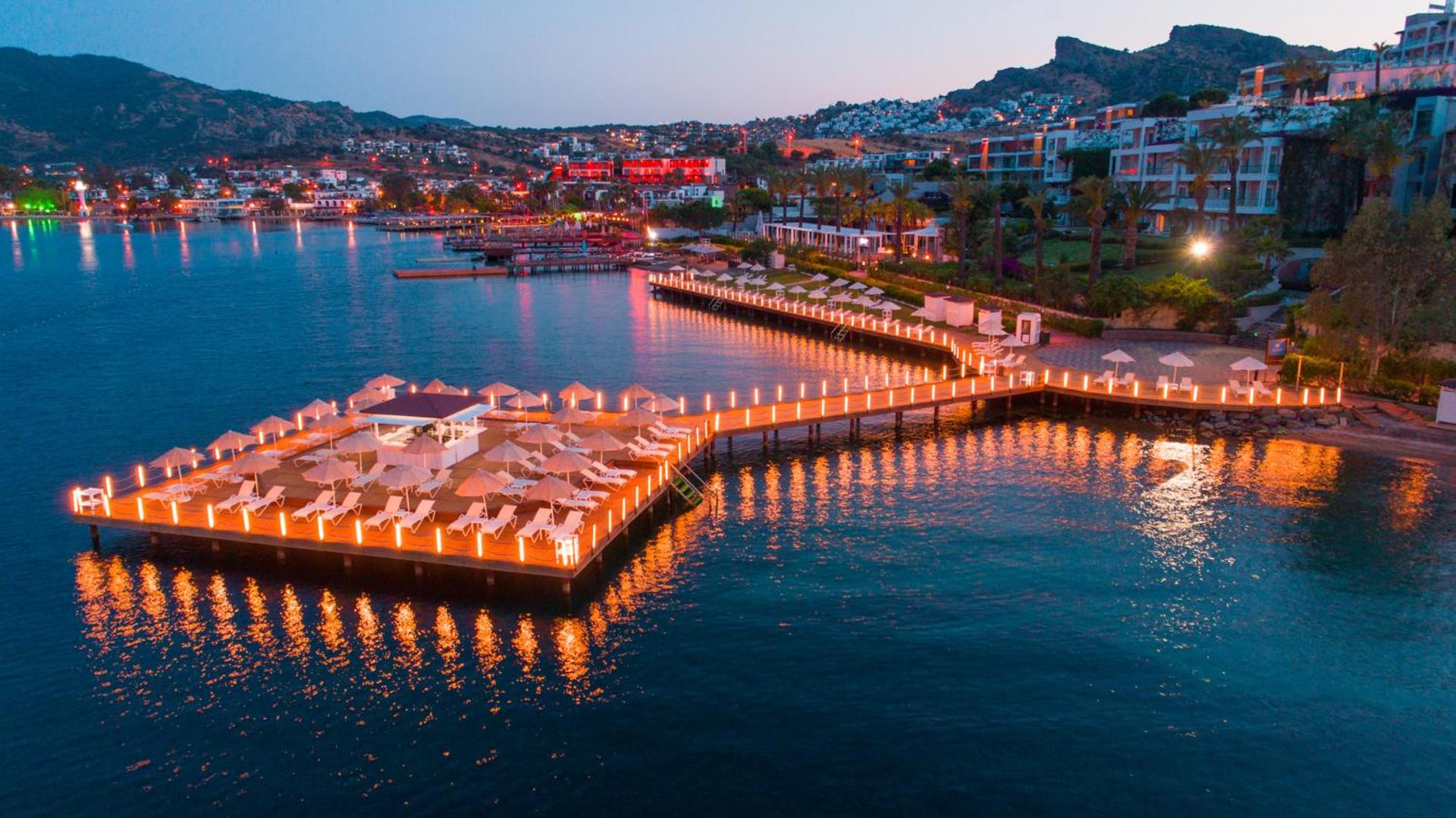 Hotel Baia Bodrum izgaismotā koka piestātne ar bāru un atpūtas krēsliem krēslas stundā Bodrumas piekrastē