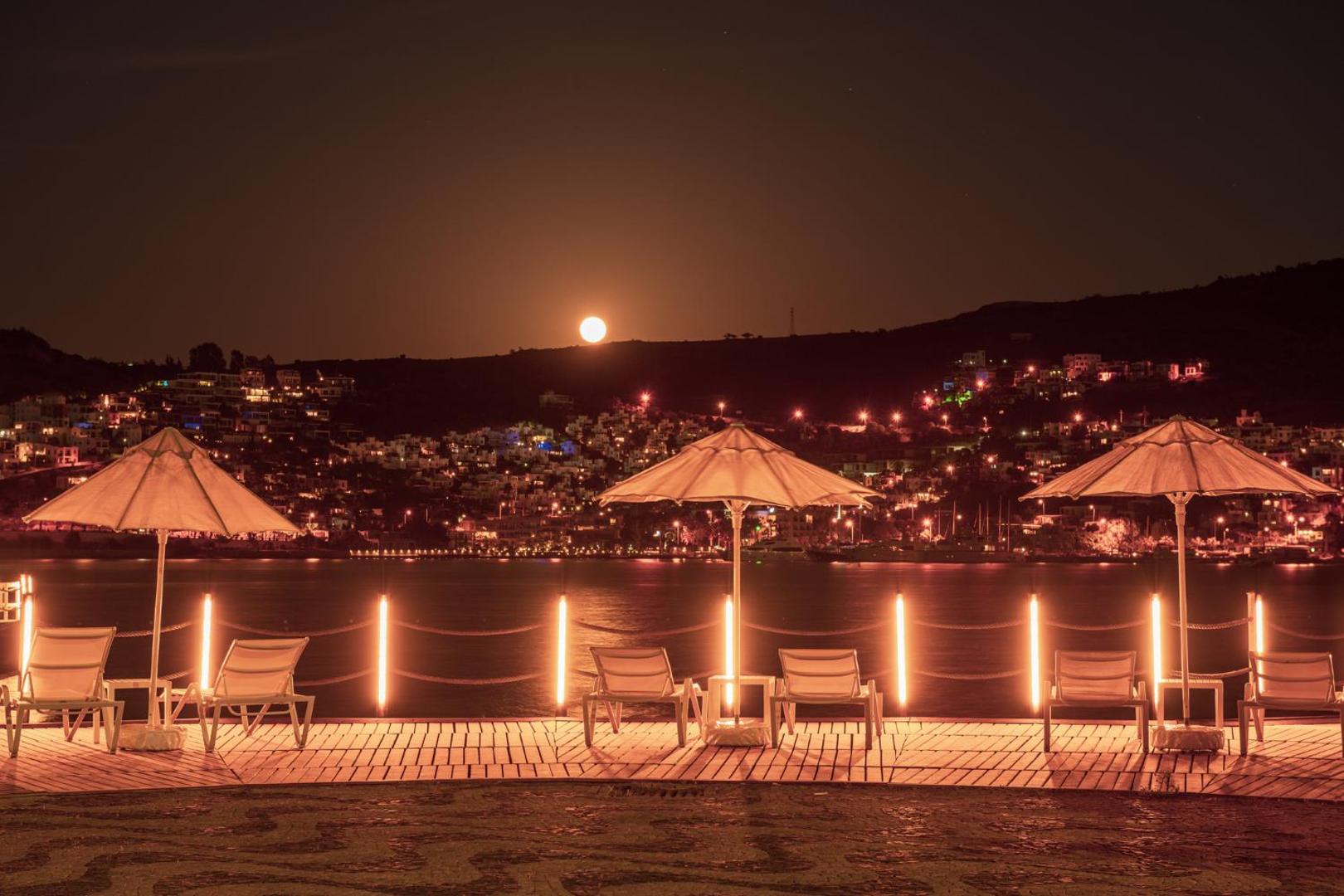 Hotel Baia Bodrum koka terase pie jūras nakts laikā ar sauļošanās krēsliem, saulessargiem un pilnmēnesi virs kalniem Bodrumas piekrastē