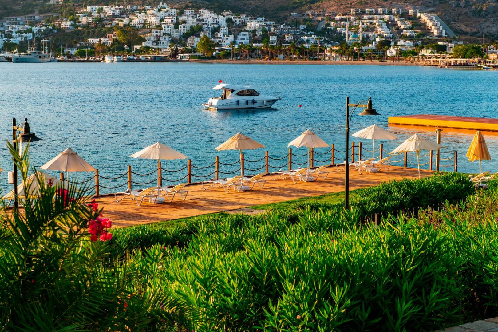 Viesnīcas Hotel Baia Bodrum koka sauļošanās terase pie jūras ar saulessargiem un skatu uz jahtu un kalniem