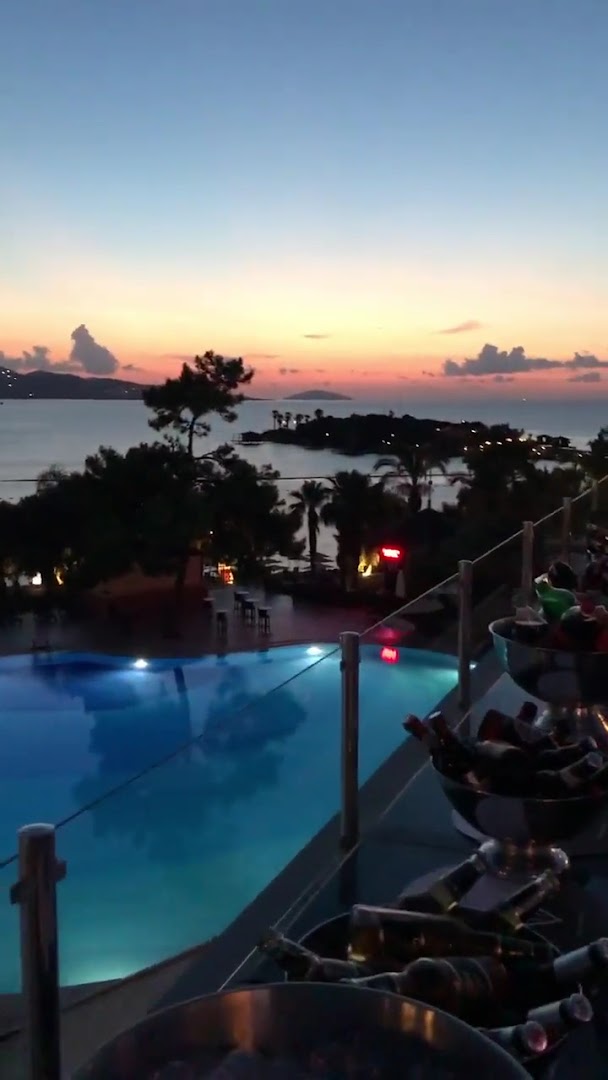 Skats no Rixos Premium Bodrum terases uz izgaismotu baseinu un jūras līci saulrieta laikā Bodrumas piekrastē