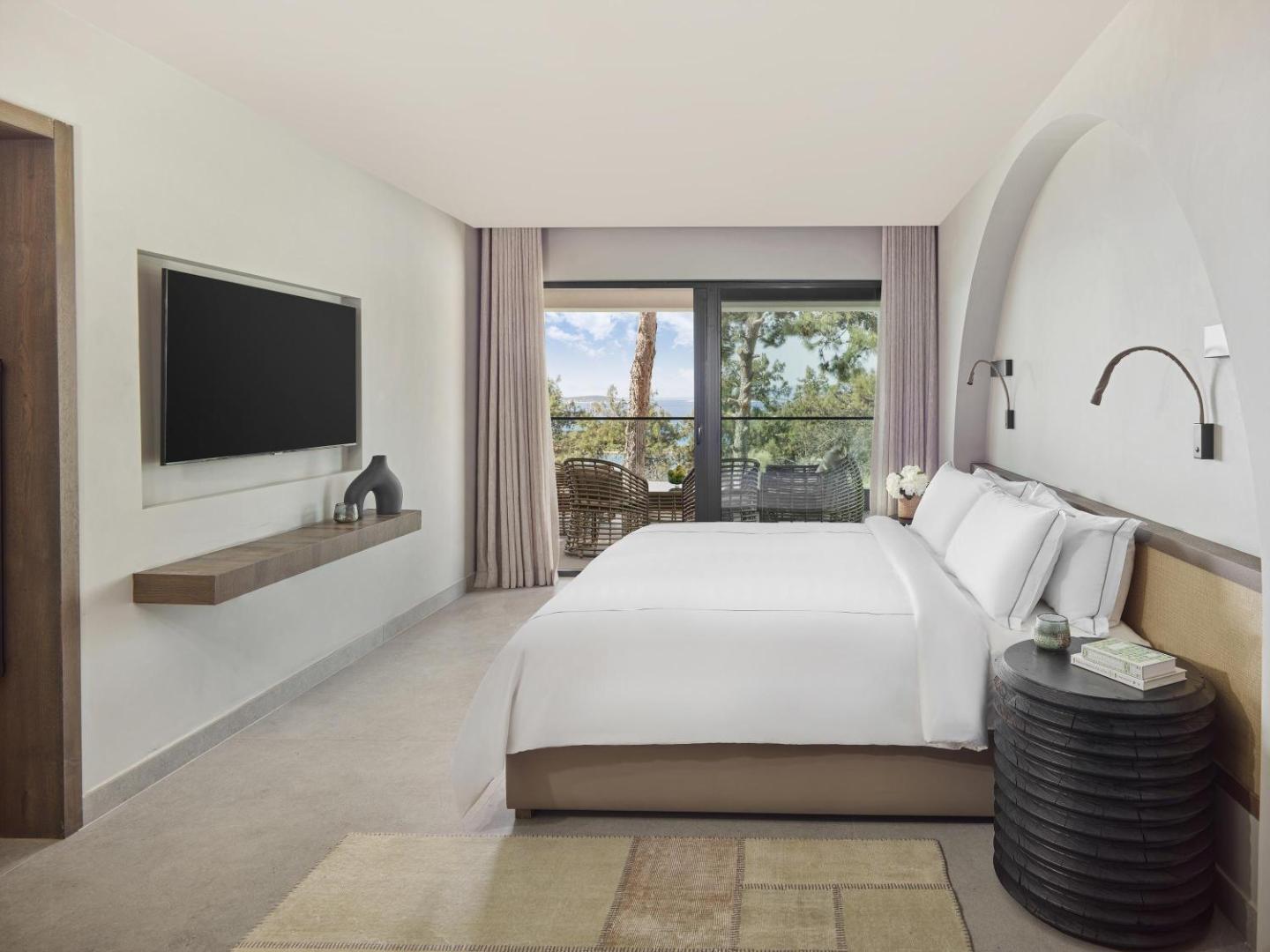 Rixos Premium Bodrum guļamistaba ar minimālistisku dizainu, sienas televizoru un skatu caur logu uz priedēm un jūru