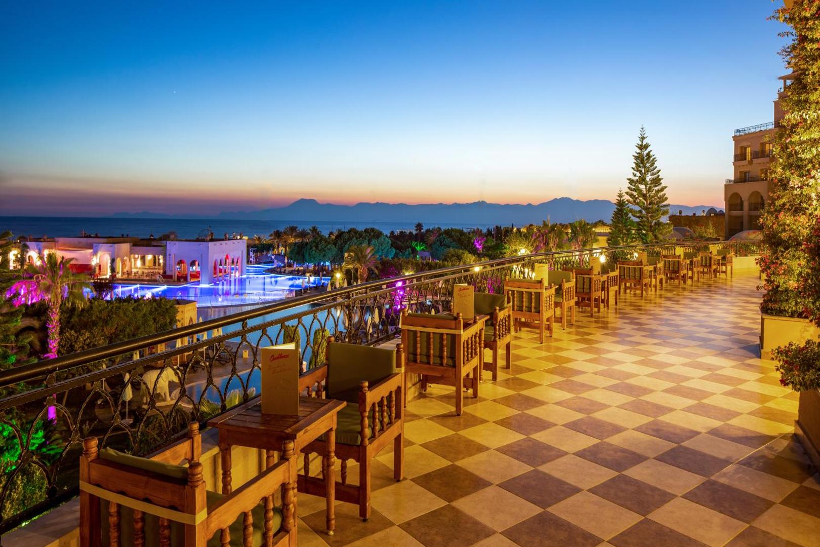 Spice Hotel & Spa plašā terase saulrietā ar skatu uz izgaismotiem baseiniem, dārzu un Vidusjūras piekrasti Belekā, Turcijā