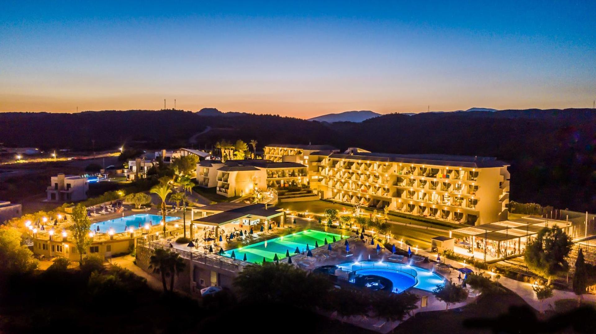 Princess Sun Hotel Rodā vakara apgaismojumā — izgaismoti baseini un viesnīcas korpusi uz kalnu un saulrieta fona