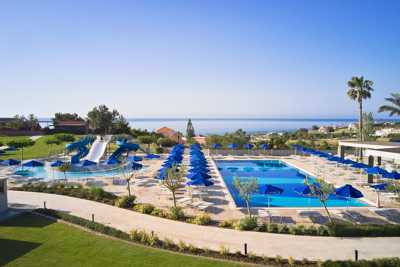 Princess Sun Hotel panorāmas skats uz baseiniem un Vidusjūru Rodas salā — plaša atpūtas zona ar zilajiem saulessargiem un ūdens slidkalniņiem