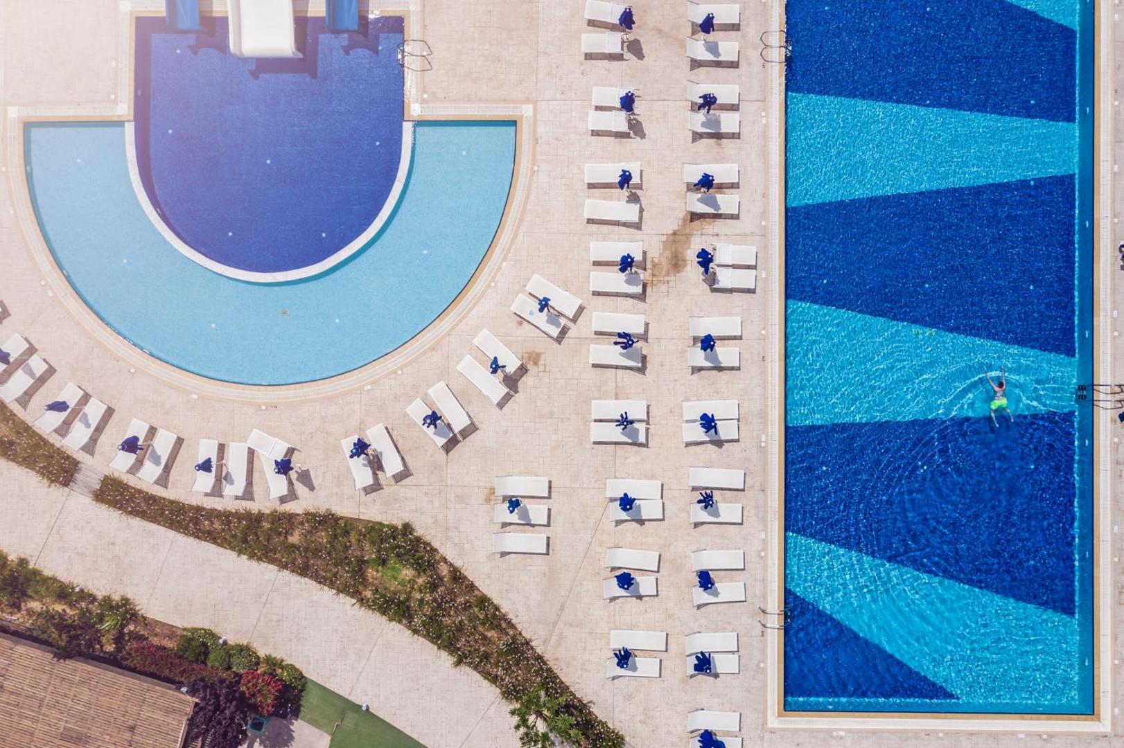 Princess Sun Hotel baseinu zonas skats no augšas Rodas salā — taisnstūra un pusapļa baseini ar sauļošanās krēsliem un saulessargiem