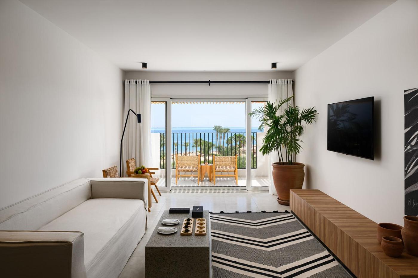 Serry Beach Resort viesistabas zona ar baltu dīvānu, koka mēbelēm un izeju uz balkonu ar skatu uz piekrasti Hurgadā.