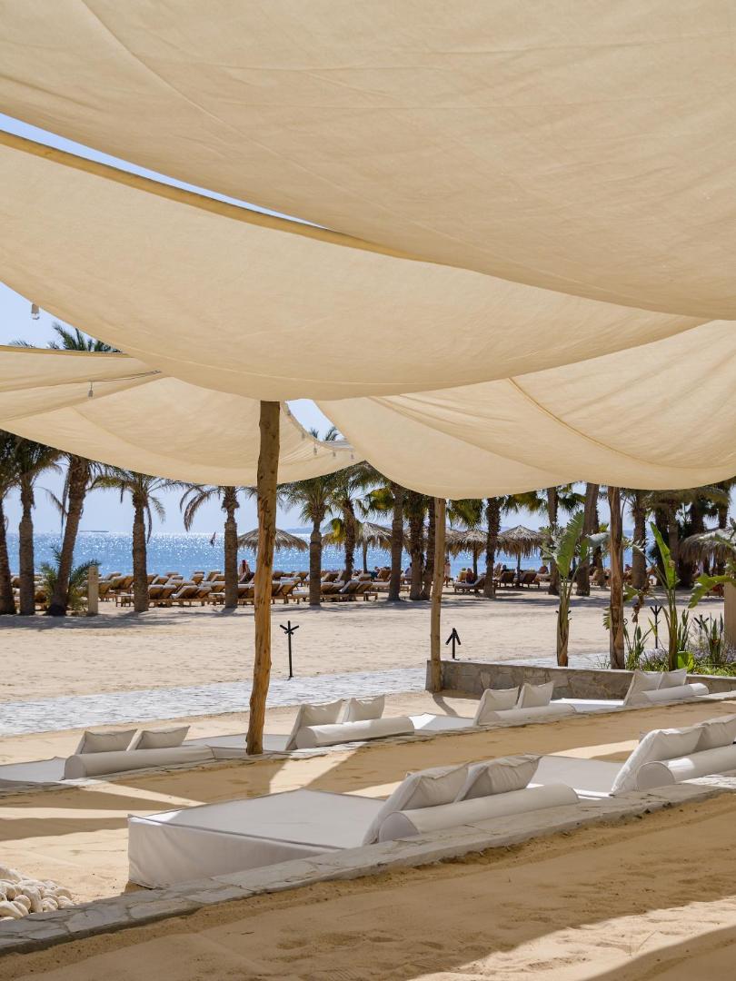 Skats no noēnotas atpūtas zonas uz Serry Beach Resort pludmali ar palmām un sauļošanās krēsliem Sarkanās jūras fonā.