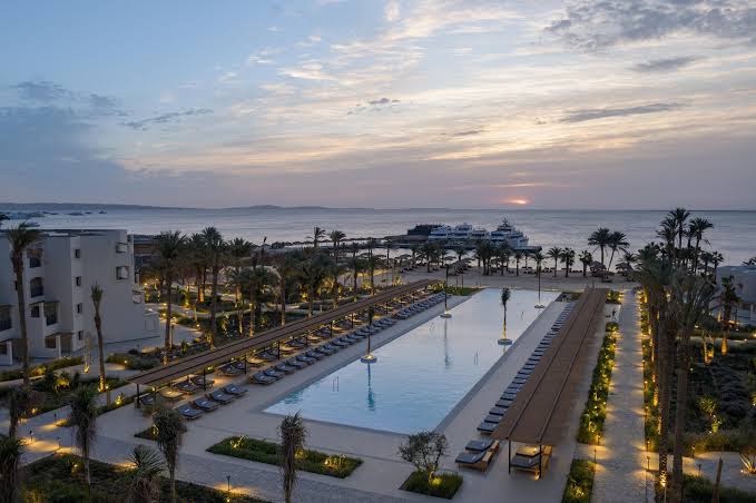 Serry Beach Resort skats no augšas uz garu āra baseinu saulrietā — apgaismoti celiņi, palmas un jūra fonā Hurgadā