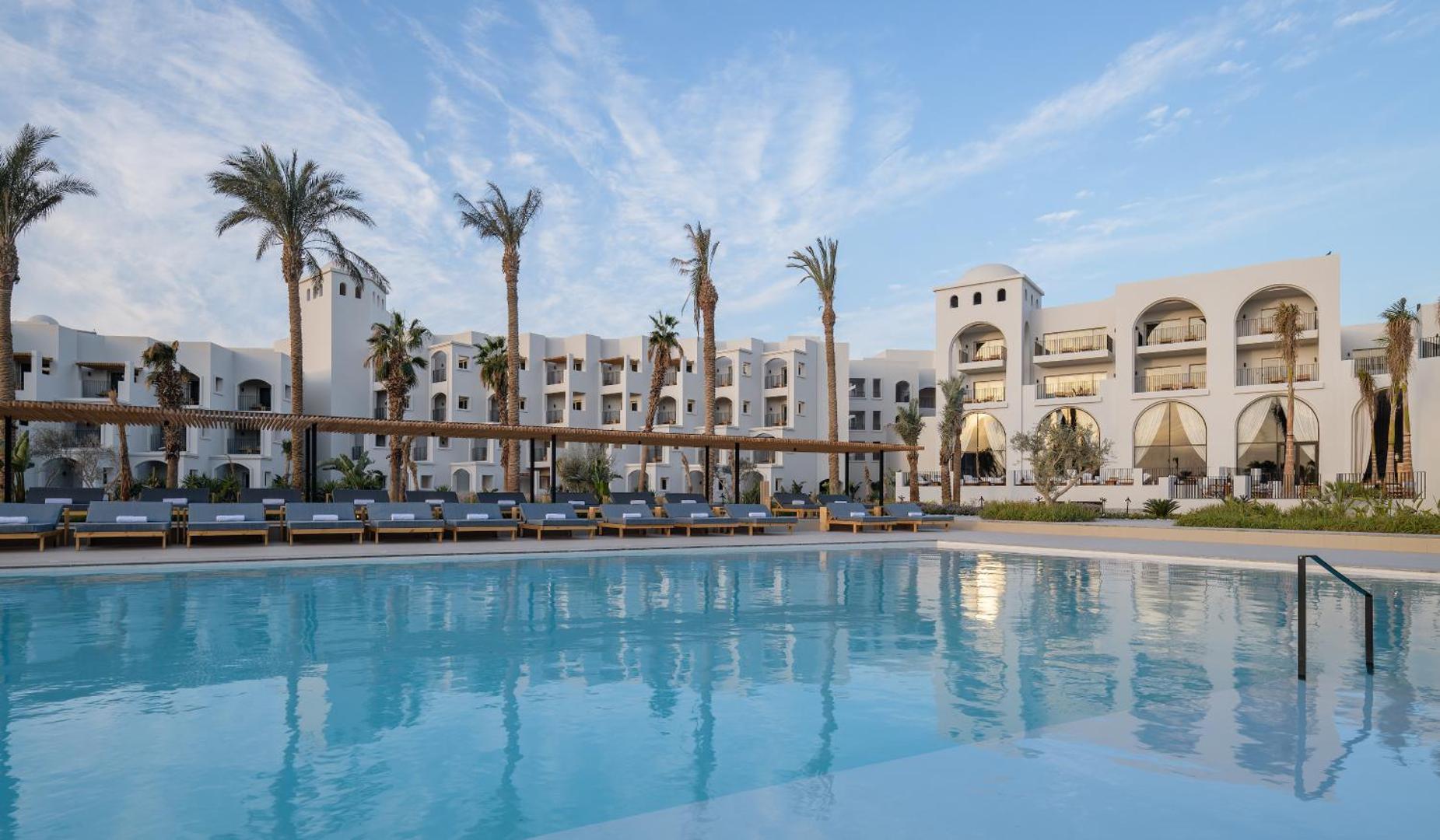 Serry Beach Resort baltie viesnīcas korpusi un baseins dienas laikā Hurgadā — moderna arhitektūra ar arkām un palmām