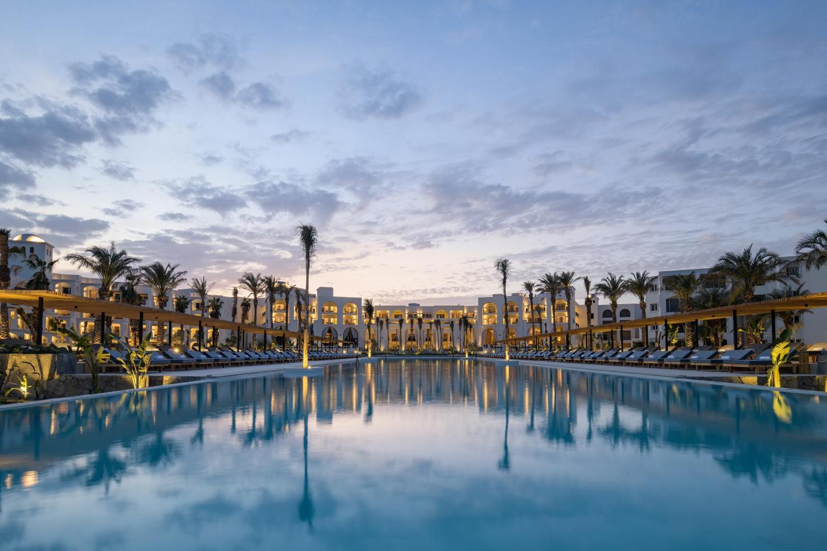 Serry Beach Resort panorāmas skats uz baseinu un viesnīcas korpusiem krēslas stundā Hurgadā — arhitektūras atspulgi ūdenī