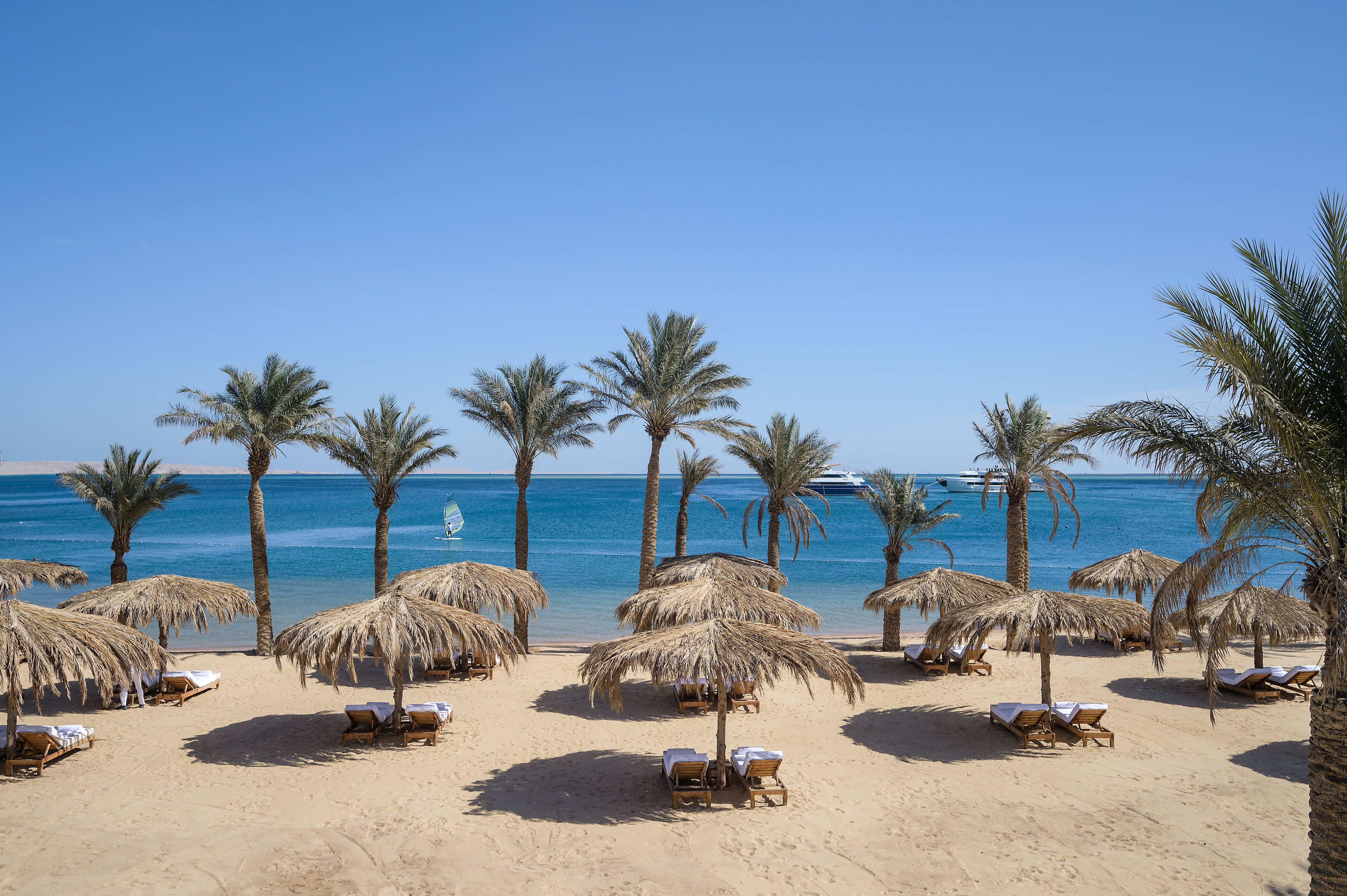 Serry Beach Resort pludmale Hurgadā ar salmu saulessargiem, koka sauļošanās krēsliem un palmām pie dzidri zilās Sarkanās jūras.