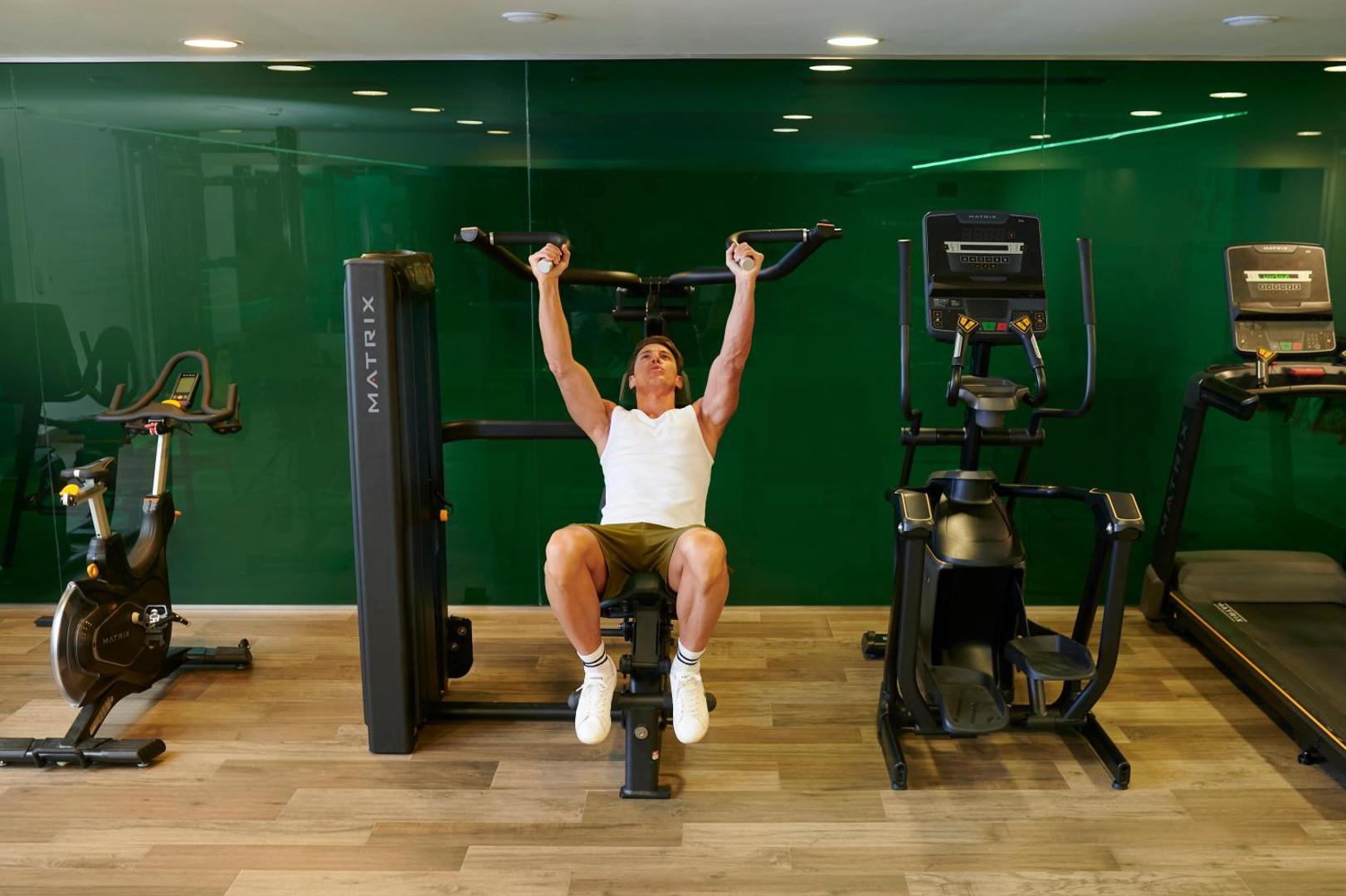 Electra Palace mūsdienīgs fitness centrs ar Matrix trenažieriem un zaļu stikla sienu — vīrietis veic spēka vingrinājumus sporta zālē