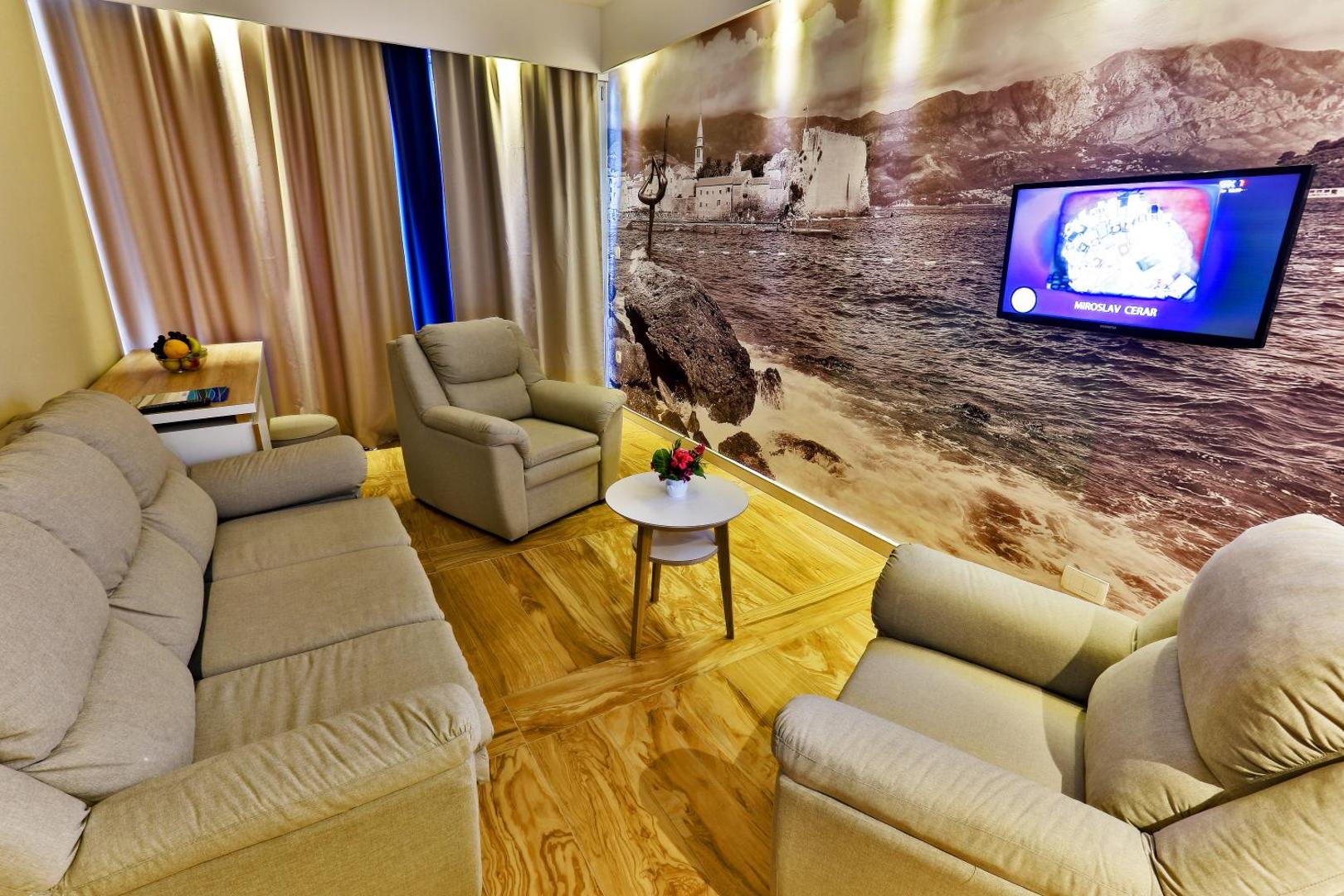 Carine Hotel Delfin viesnīcas numura atpūtas zona ar dīvānu, atpūtas krēsliem, televizoru un dekoratīvu sienas attēlu ar piekrastes ainavu