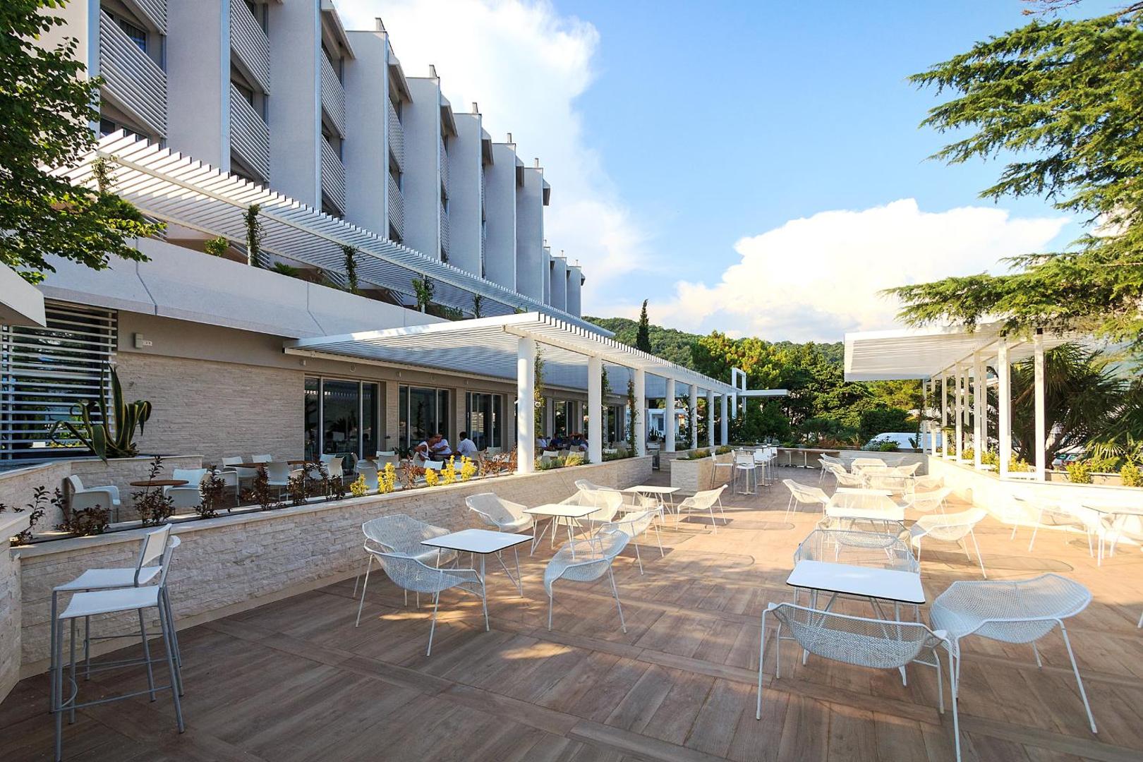 Carine Hotel Delfin āra terase ar modernām mēbelēm un saulessargiem pie viesnīcas fasādes Melnkalnē, skats uz kalniem