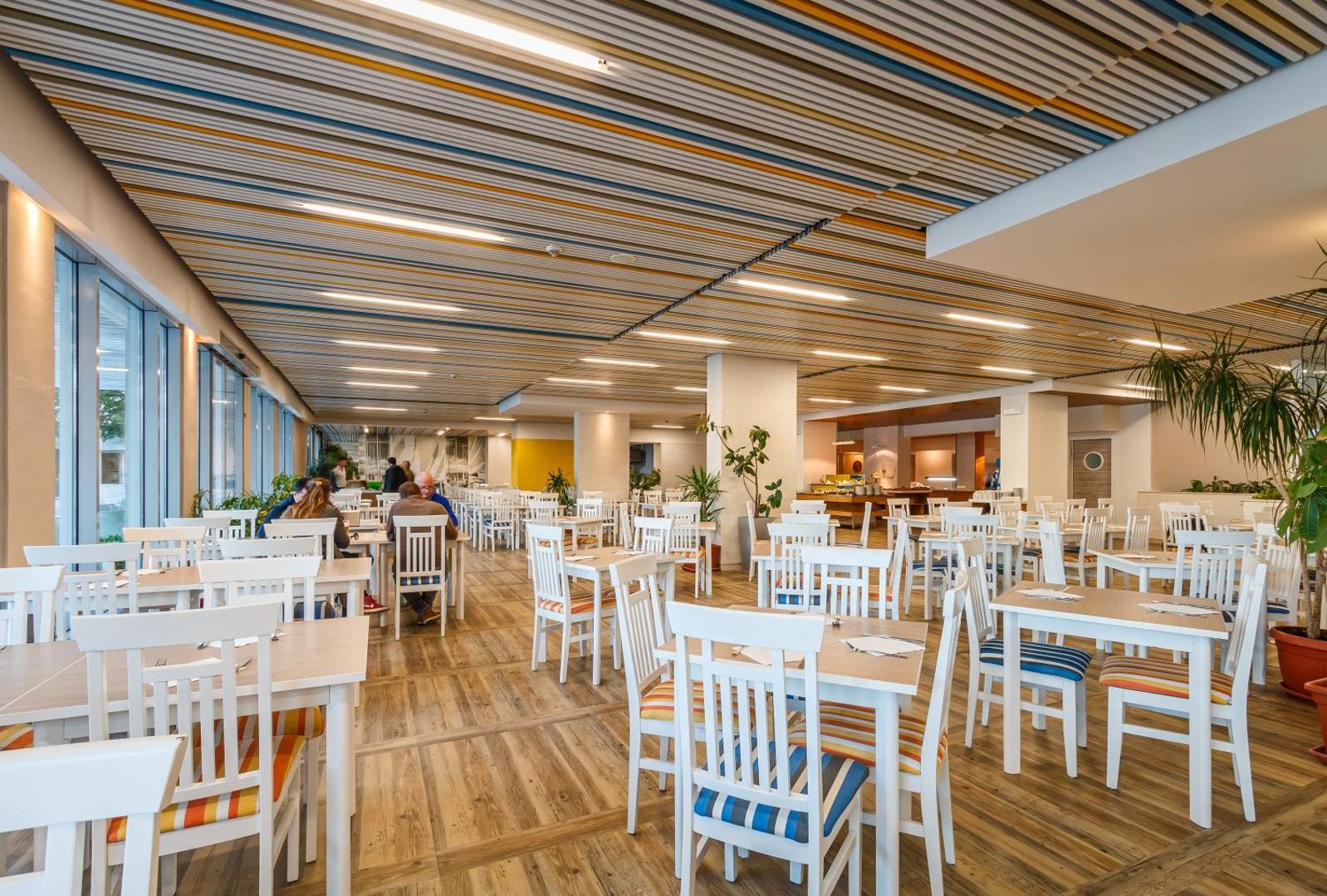 Carine Hotel Delfin plašais restorāns ar modernu interjeru, gaišām mēbelēm un lieliem logiem uz jūras pusi