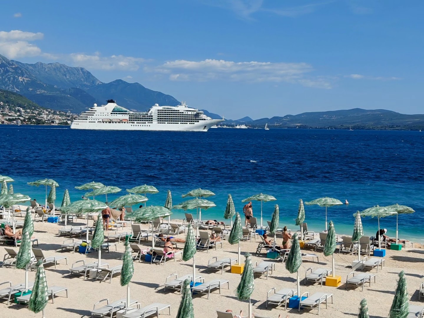 Skats no Carine Hotel Delfin pludmales uz zilo jūru, kurā kuģo liels kruīza kuģis uz kalnu fona Melnkalnē