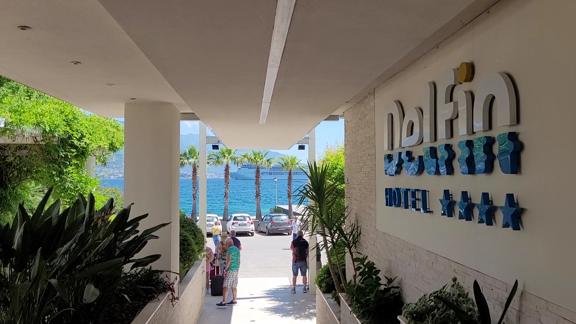 Ieeja Carine Hotel Delfin ar skatu uz jūru un kruīza kuģi tālumā — viesnīcas 4* zīme pie sienas