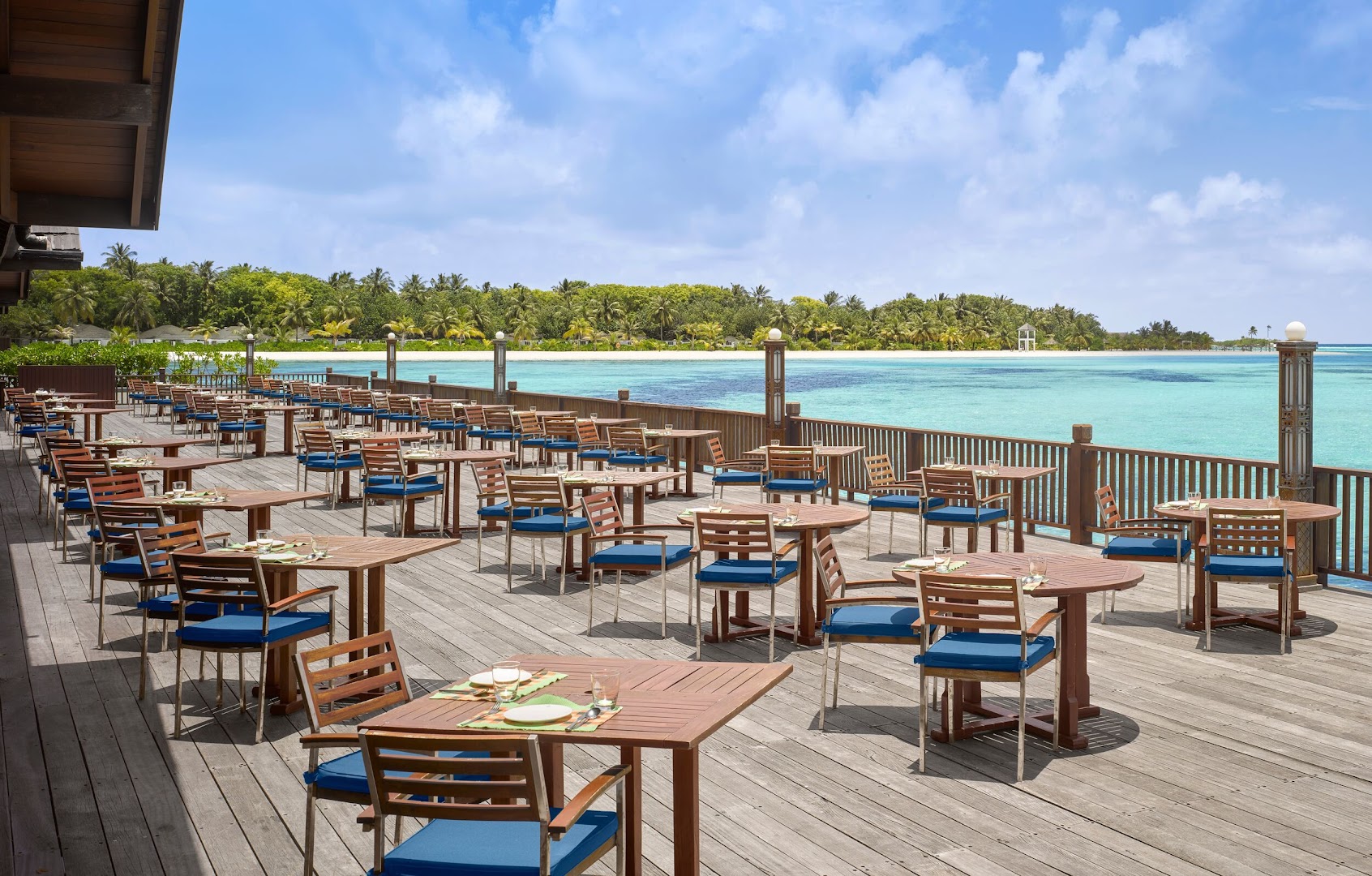 Villa Nautica Paradise Island restorāna āra terase ar koka galdiem un zilām mēbelēm pie okeāna Maldīvu salās