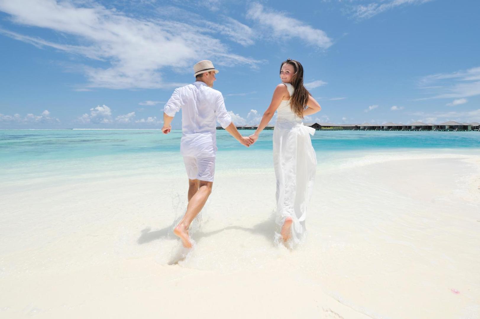 Jaunlaulātie baltās drēbēs skrien pa seklu okeāna ūdeni Villa Nautica Paradise Island pludmalē — fonā ūdens bungalo un zilas debesis