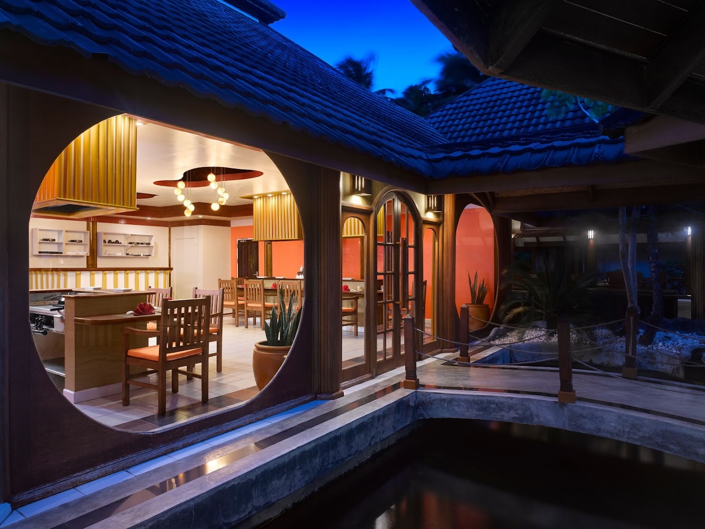 Villa Nautica Paradise Island restorāna interjers ar apaļu logu un skatu uz dīķi naktī — elegants apgaismojums un koka apdare