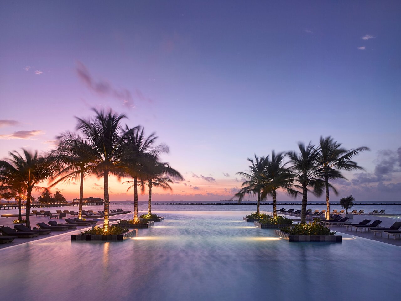 Villa Nautica Paradise Island bezgalības baseins saulrietā ar palmām un izgaismotu atpūtas zonu pie okeāna Maldīvu salās