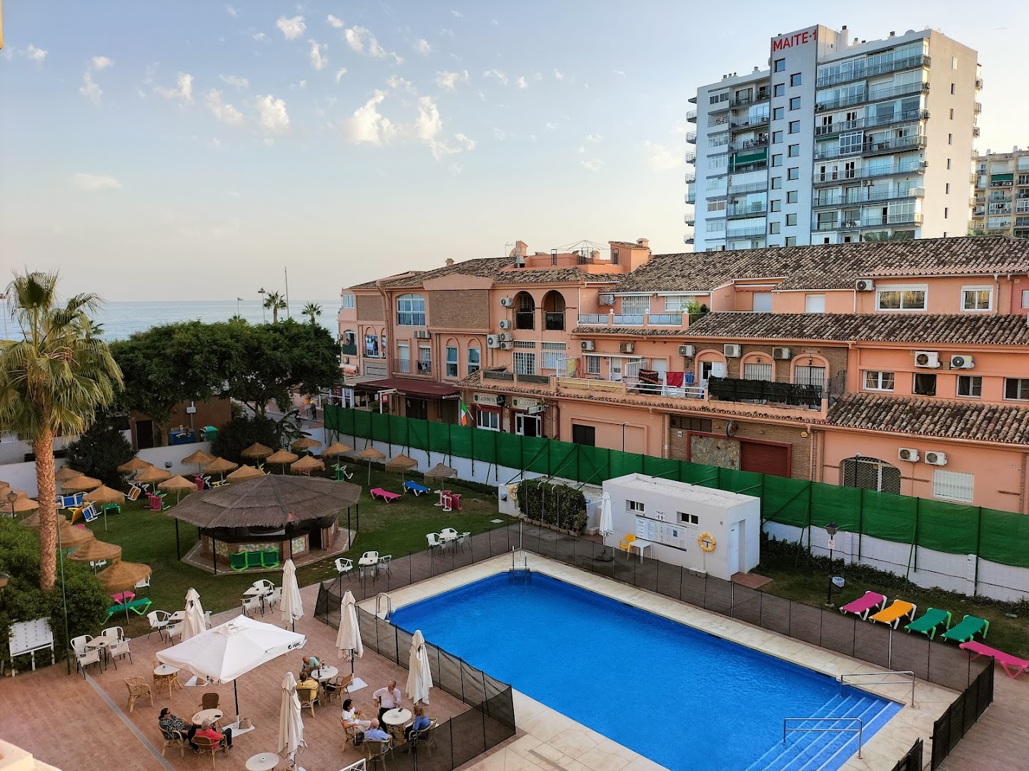 MedPlaya Hotel Alba Beach baseina zona vakarā ar galdiņiem un saulessargiem — fonā Benalmadenas pilsētas apbūve un jūra saulrietā