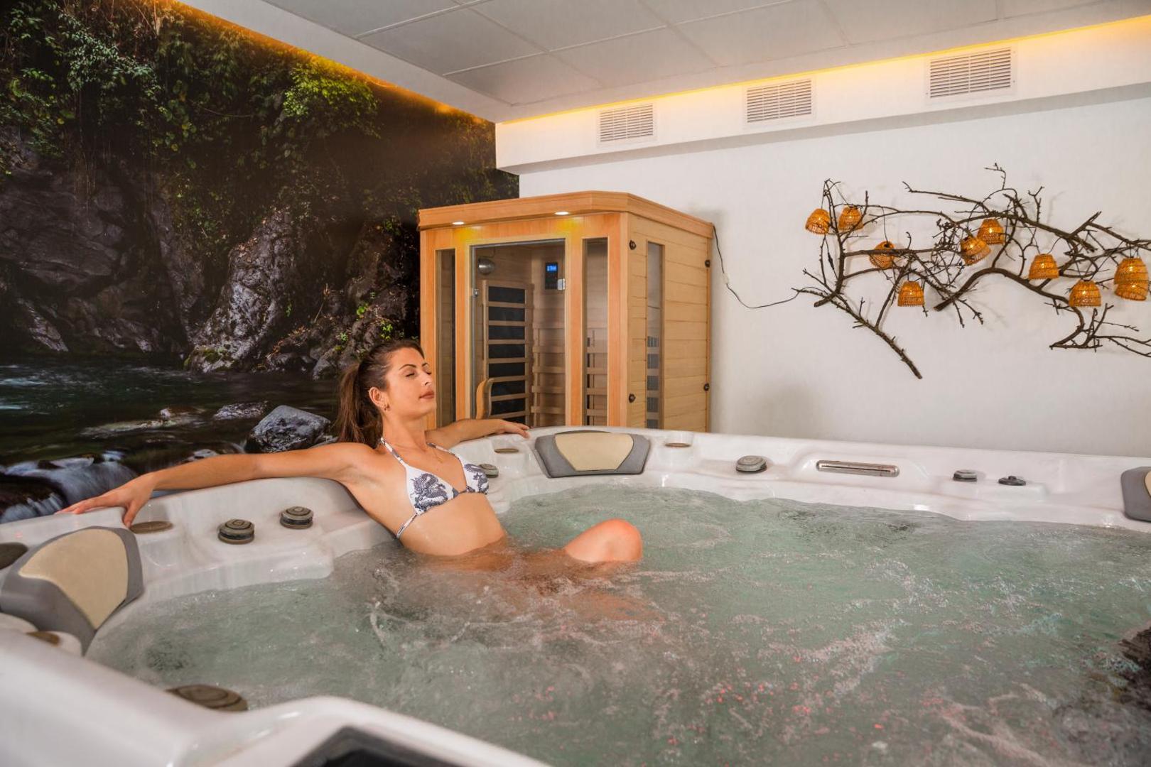Hotel Alay spa zona ar lielu džakuzi vannu un sievieti tajā — fonā dabas motīvu sienas gleznojums un neliela sauna