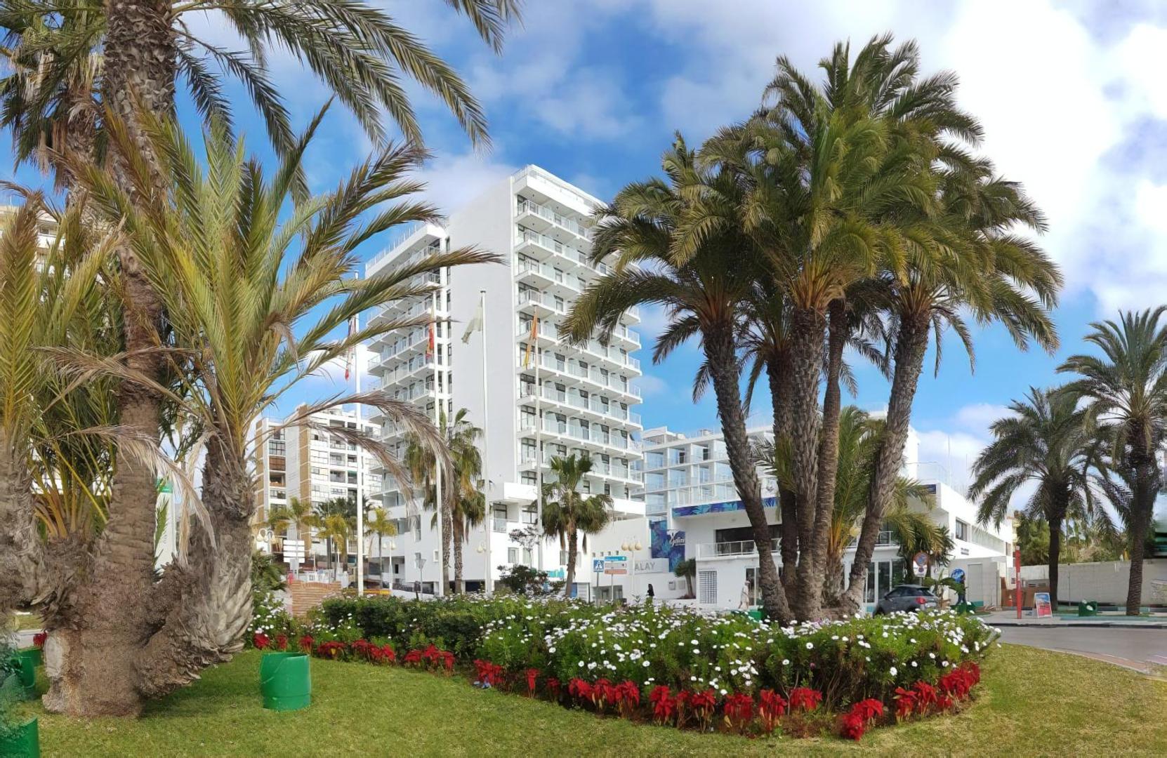 Viesnīcas Hotel Alay modernā daudzstāvu ēka Benalmadenā, Spānijā — skats no dārza puses ar palmām un ziedošām puķu dobēm