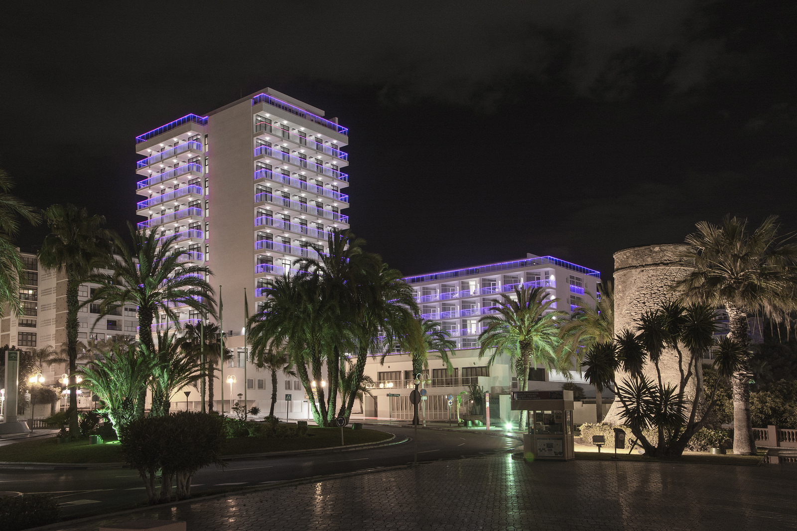 Hotel Alay nakts skats Benalmadenā — izgaismota viesnīcas fasāde ar violetām gaismām, apkārtnē palmas un vēsturisks sargtornis