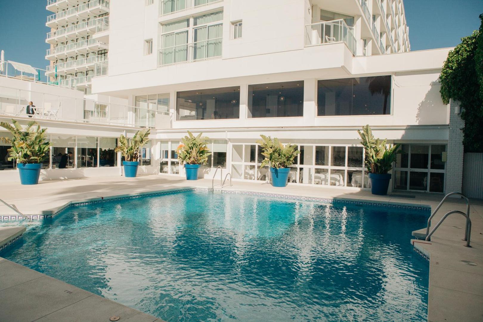 Hotel Alay iekšpagalma baseins pie baltas viesnīcas ēkas ar dekoratīviem augiem zilos podos — mājīga un saulaina peldvieta
