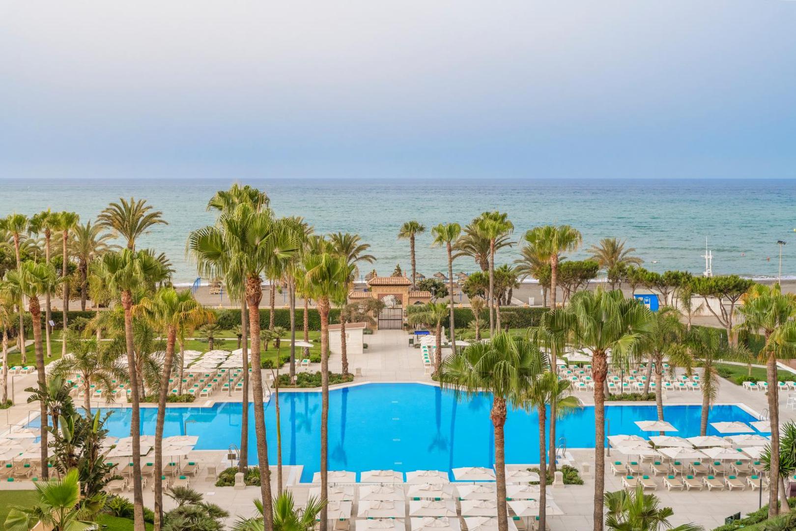 Skats no augšas uz Iberostar Malaga Playa āra baseinu, palmu dārzu un Vidusjūras piekrasti — atpūtas zona ar saulessargiem pie pludmales
