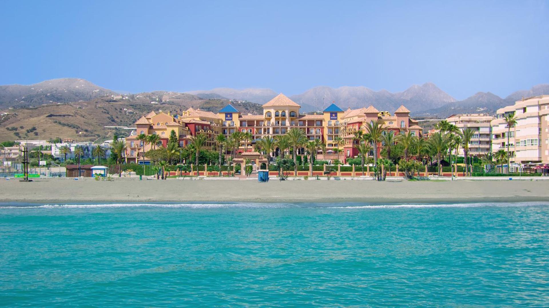 Skats no jūras uz Iberostar Malaga Playa viesnīcu, smilšu pludmali un kalnu grēdu tālumā saulainā dienā