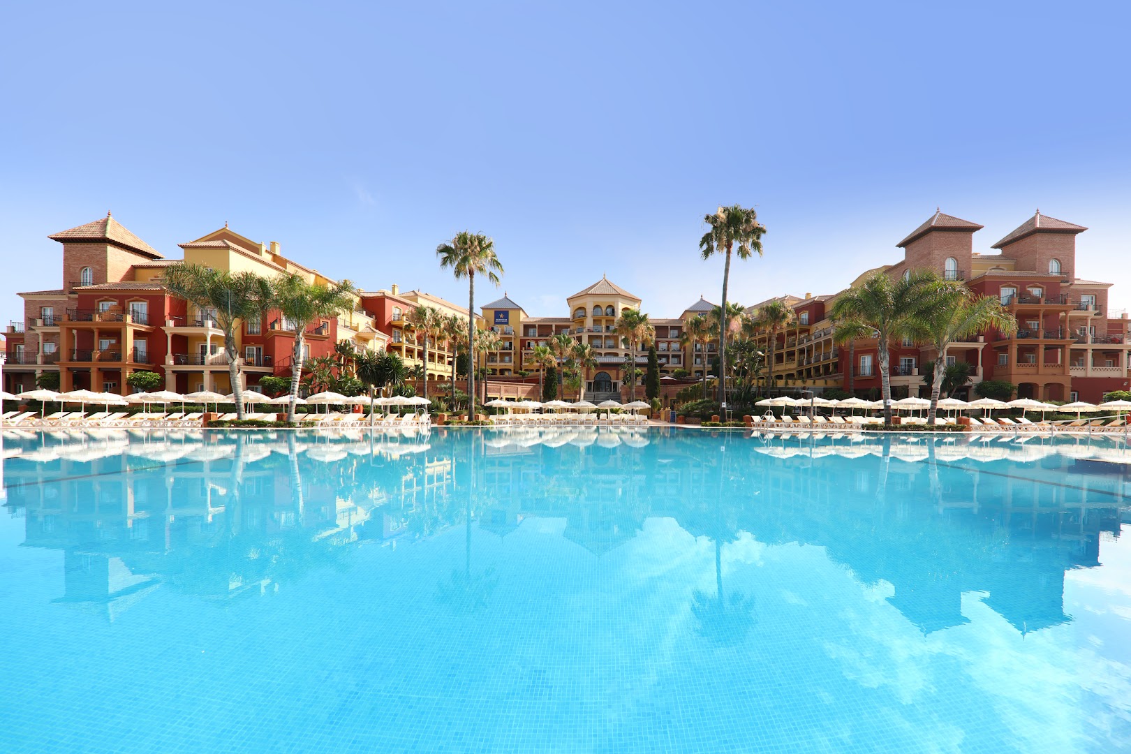 Iberostar Malaga Playa viesnīcas arhitektūra un lielais āra baseins saulainā dienā — kūrorts ar palmām un atpūtas krēsliem Andalūzijas piekrastē