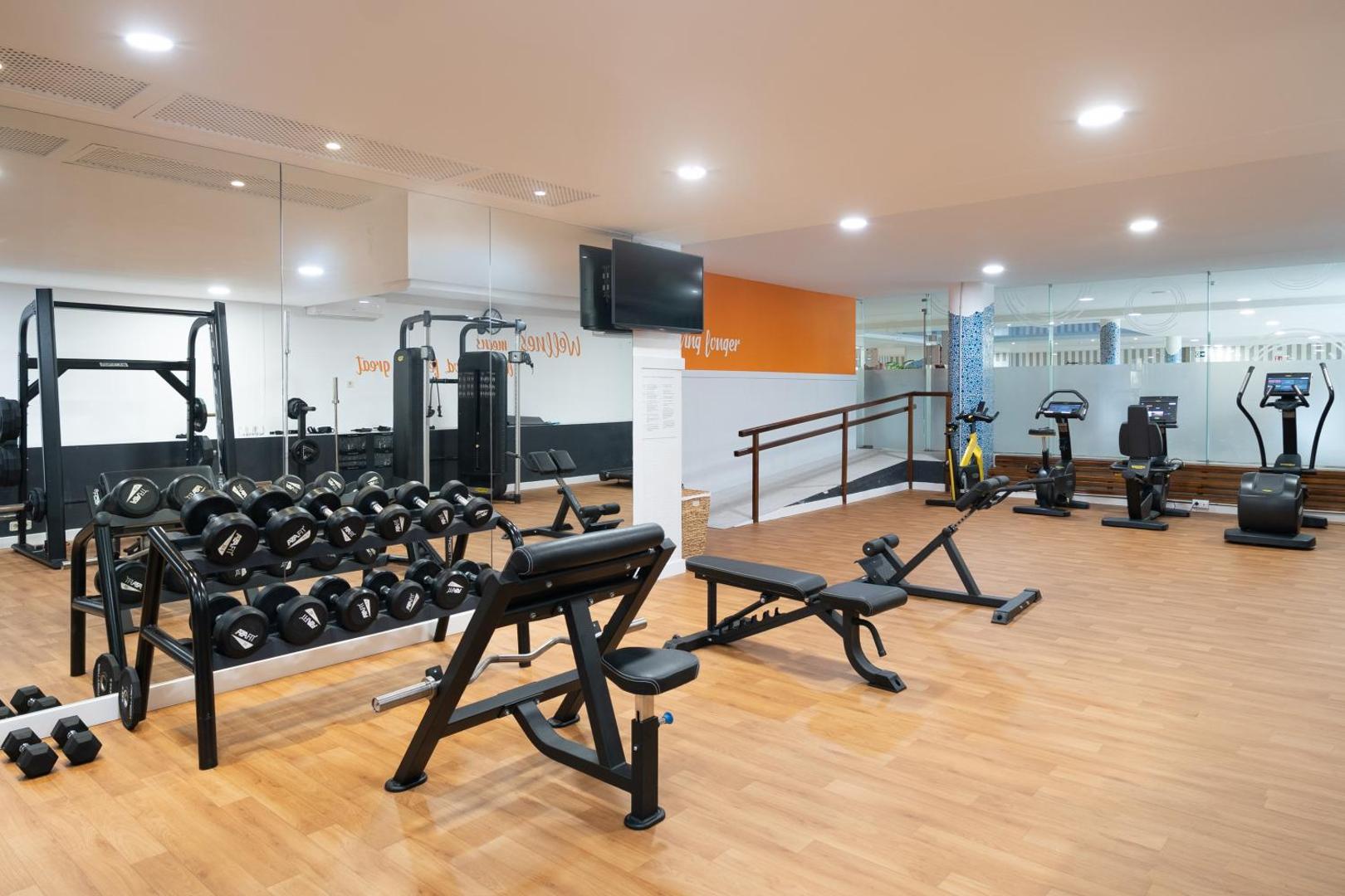 Mūsdienīgs Iberostar Malaga Playa fitness centrs ar hantelēm, skrejceliņiem un velotrenažieriem gaišā telpā ar koka grīdu.