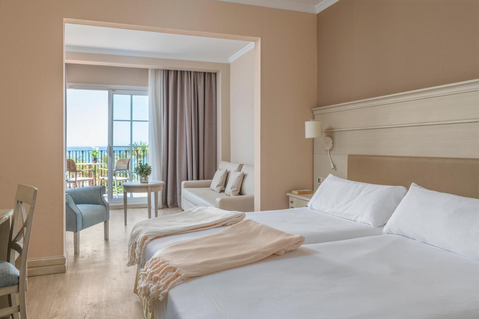 Iberostar Malaga Playa gaišs viesnīcas numurs ar divām gultām, atpūtas zonu ar dīvānu un izeju uz balkonu.