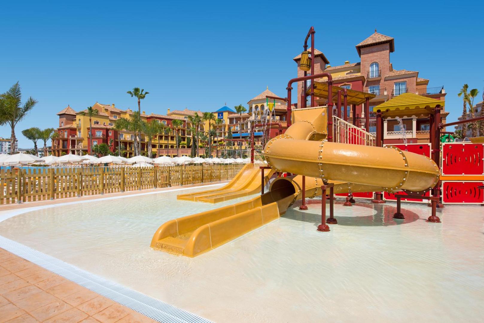 Iberostar Malaga Playa bērnu ūdens atrakciju parks ar dzelteniem slidkalniņiem un seklu baseinu uz viesnīcas ēku fona.