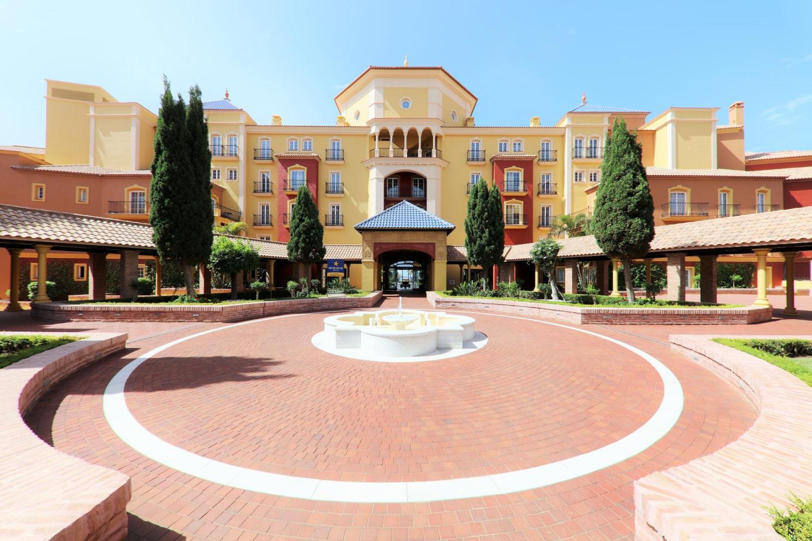 Iberostar Malaga Playa viesnīcas ieeja ar strūklaku un klasisku arhitektūru siltos toņos, ko ieskauj kopti ciprešu koki