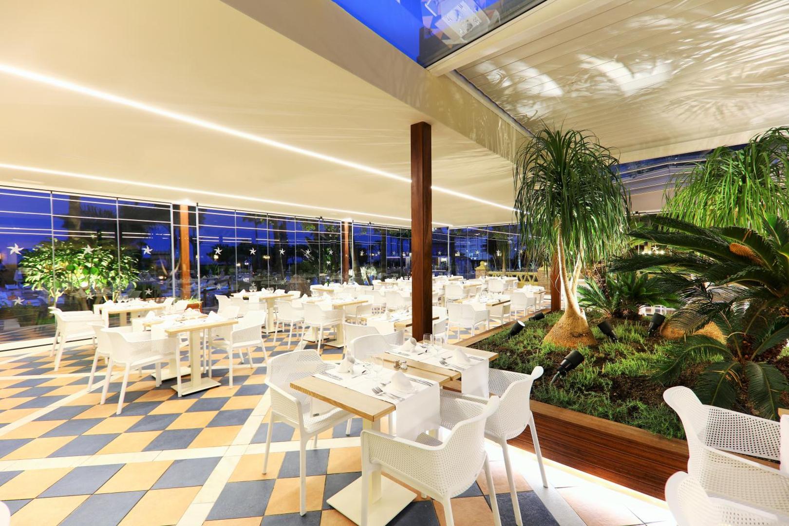 Iberostar Malaga Playa viesnīcas modernais iekštelpu restorāns ar dekoratīviem augiem un stikla sienām vakarā