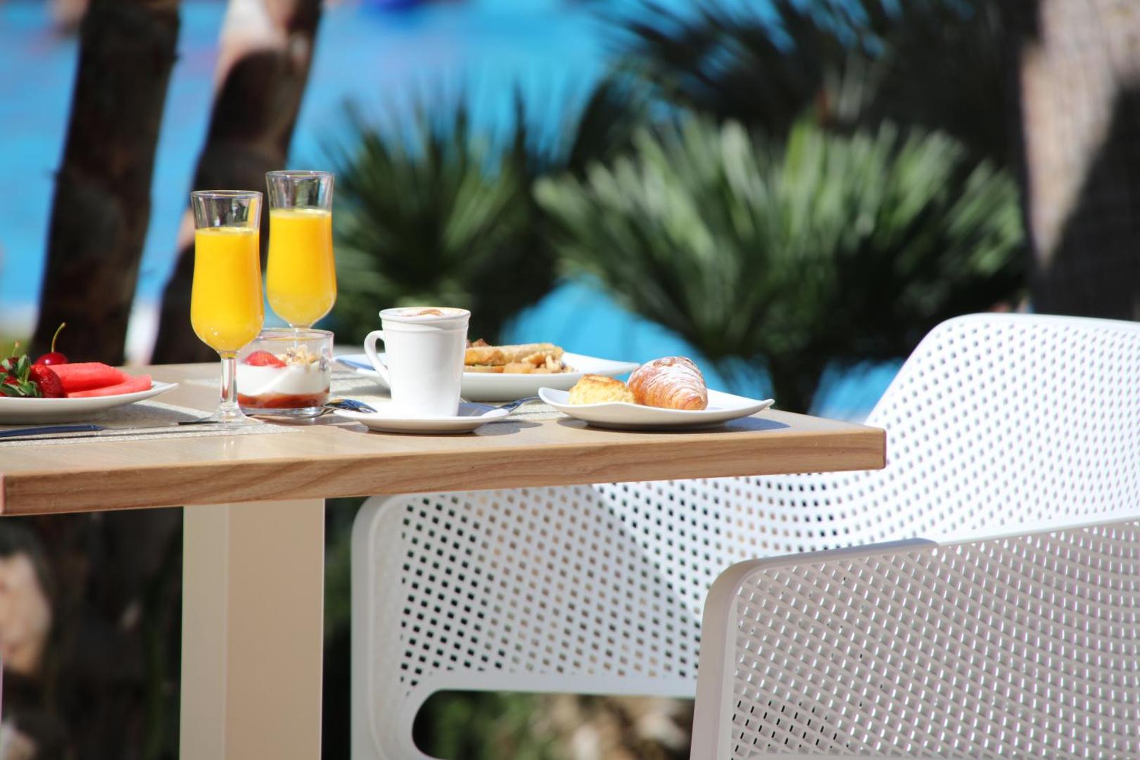Brokastu galds Iberostar Malaga Playa terasē ar svaigi spiestu apelsīnu sulu, kafiju, kruasāniem un augļiem uz zila baseina fona.